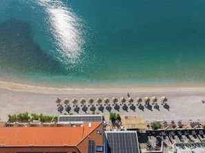 An aerial view of a beach with a row of sun loungers and a building with a red roof in the foreground. - Hotel Dobrovit (Baska)
