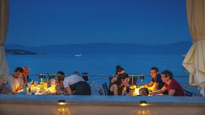 Un grupo de personas cenando en una mesa al aire libre con vistas al mar y montañas de fondo.