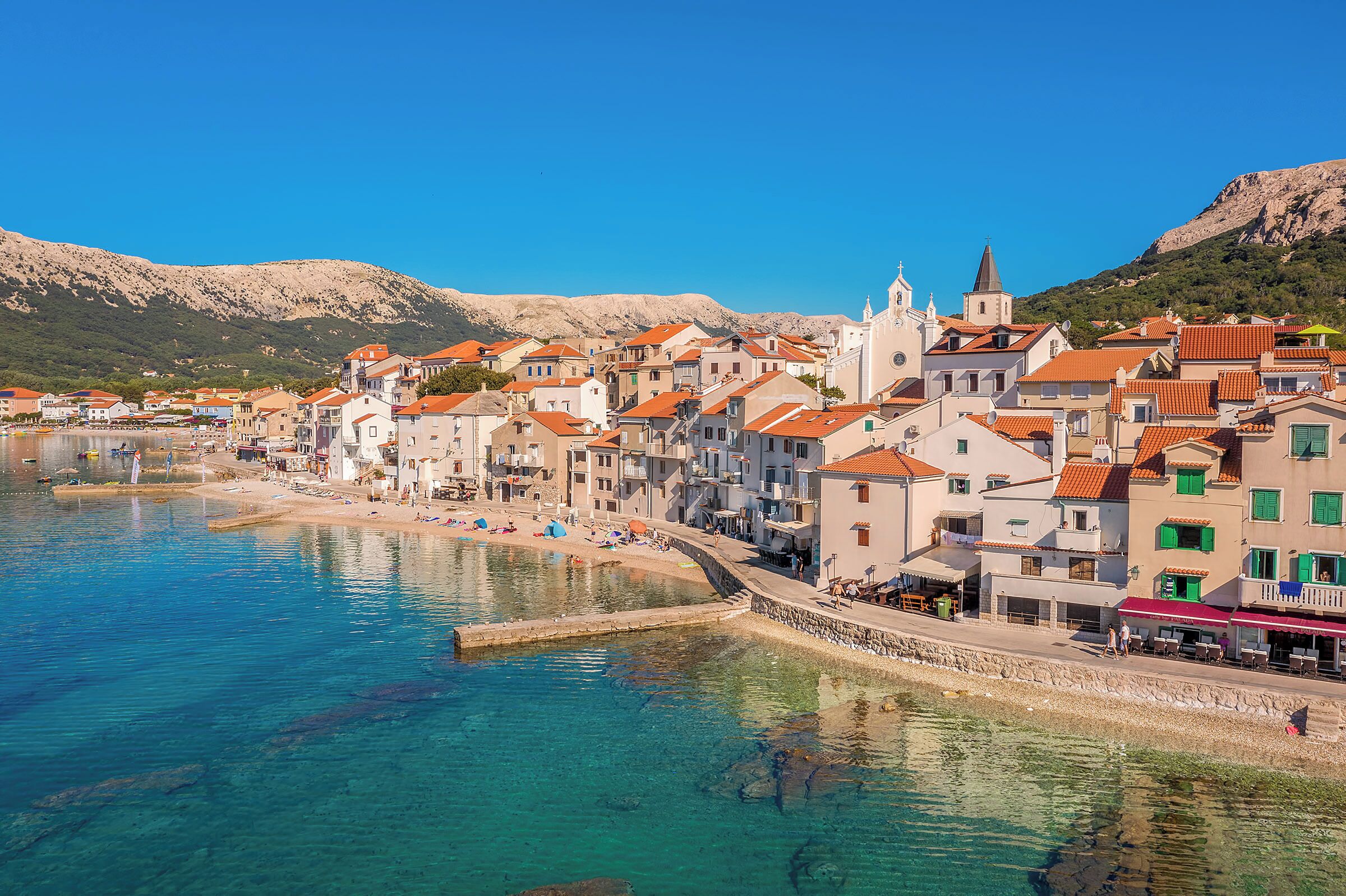 A coastal town with buildings along the waterfront, clear blue water, and a mountainous backdrop.