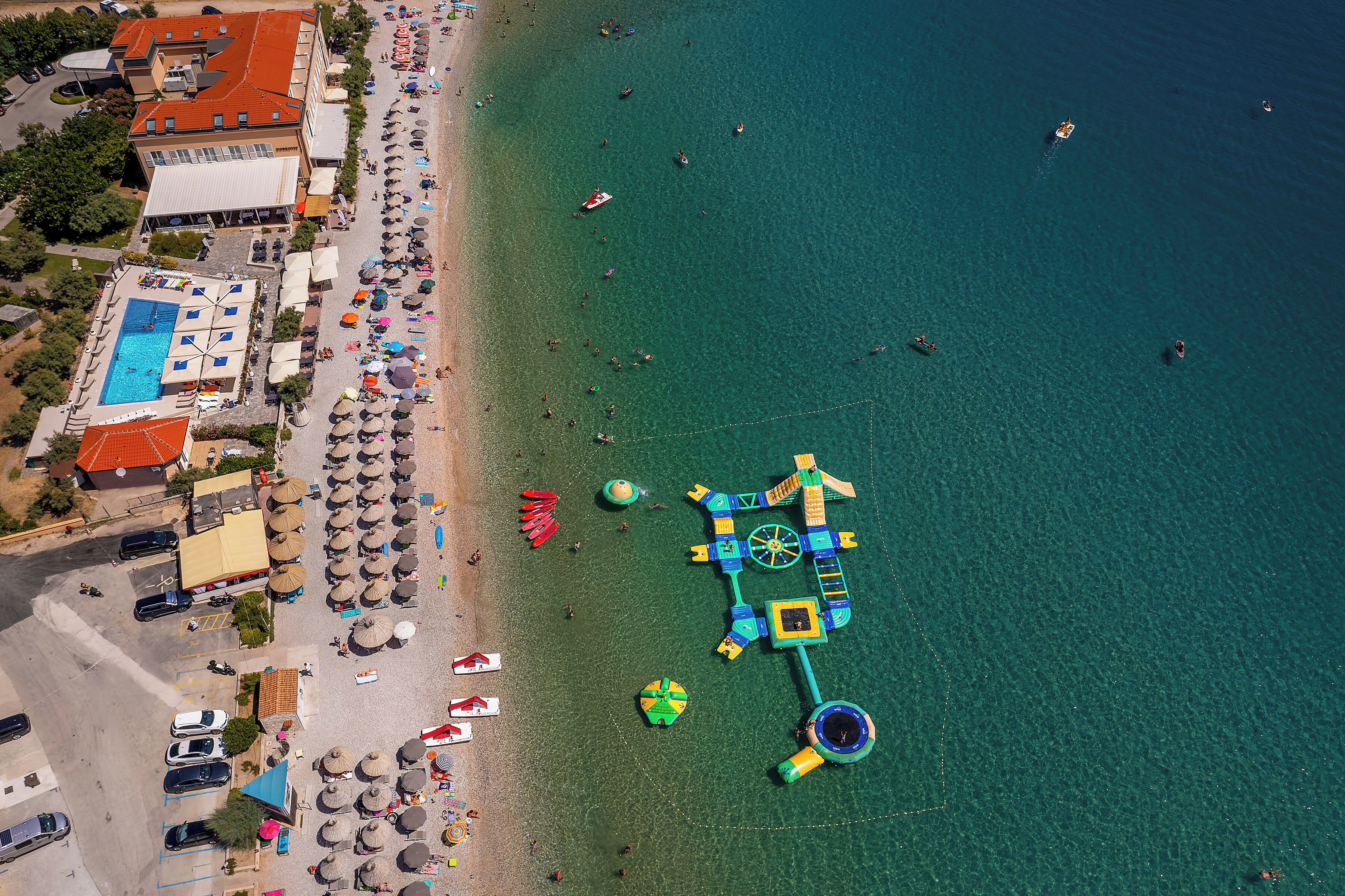 Luftaufnahme eines Strandes mit Wasserpark, einschließlich Pool, Sonnenschirmen und einem Gebäude mit rotem Dach.