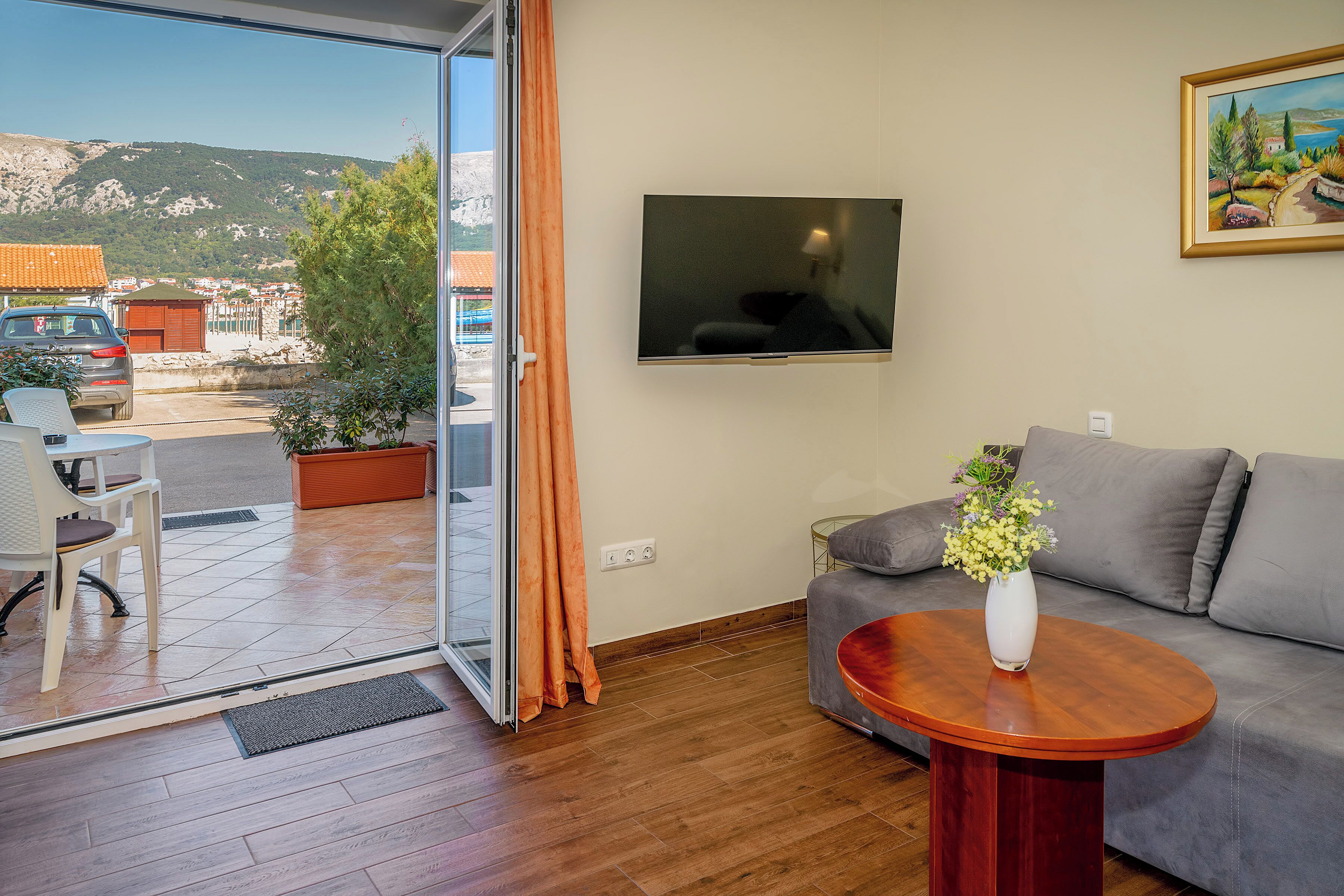 Ein modernes Wohnzimmer mit grauem Sofa, einem Holztisch, einem an der Wand befestigten Fernseher und Blick auf eine gebirgige Landschaft durch eine Glastür.