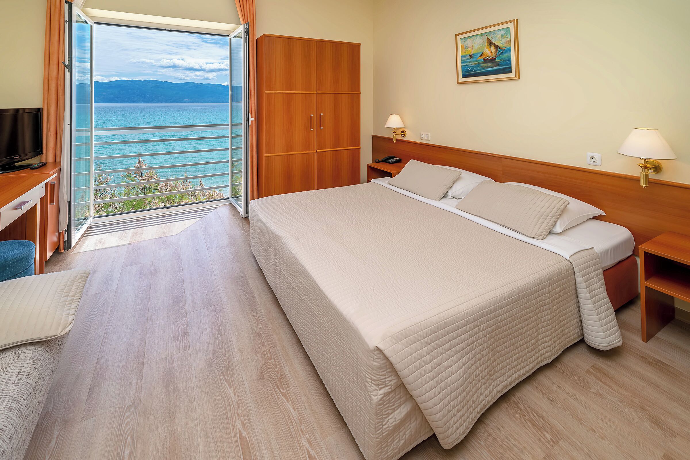 Habitación de hotel con una cama grande, un armario de madera, un escritorio con televisor y un balcón con vistas al mar.