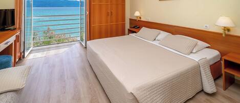 Habitación de hotel con una cama grande, un armario de madera, un escritorio con televisor y un balcón con vistas al mar.