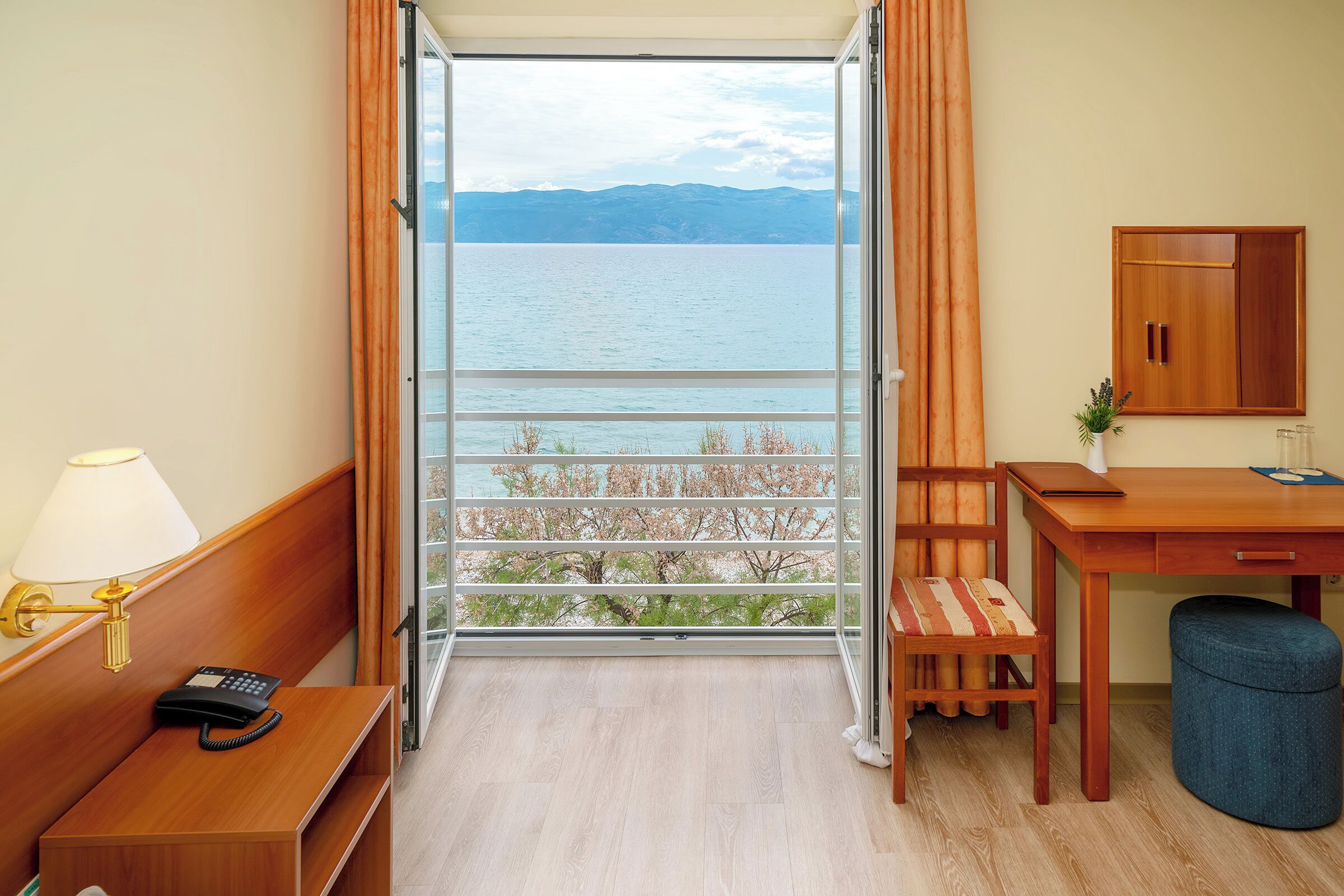 Une chambre d’hôtel avec un bureau en bois, une chaise, une petite table, une lampe, un téléphone et une vue sur un lac, visible à travers une porte vitrée.