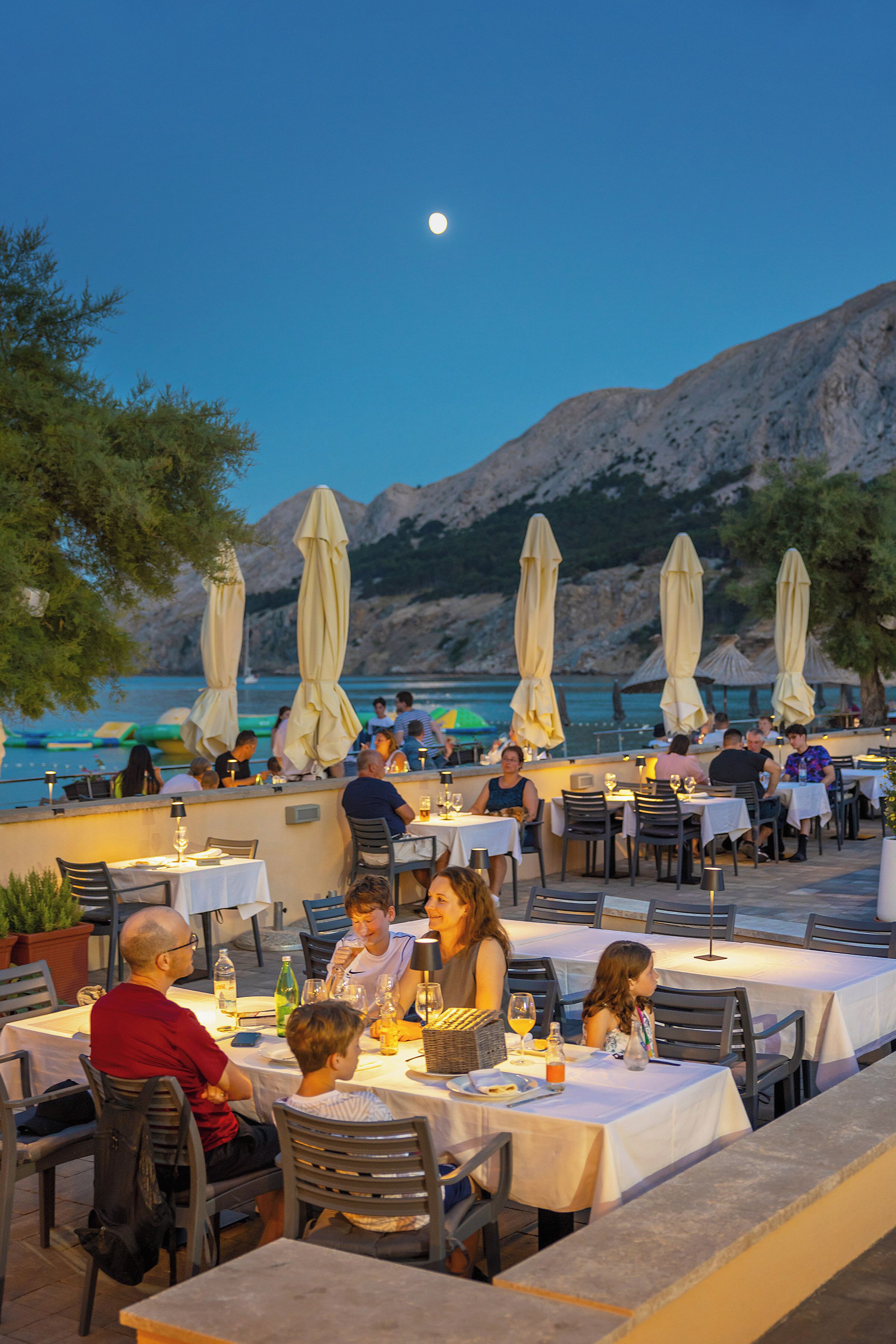 Een openluchtrestaurant met tafels en stoelen, parasols en uitzicht op zee en bergen.