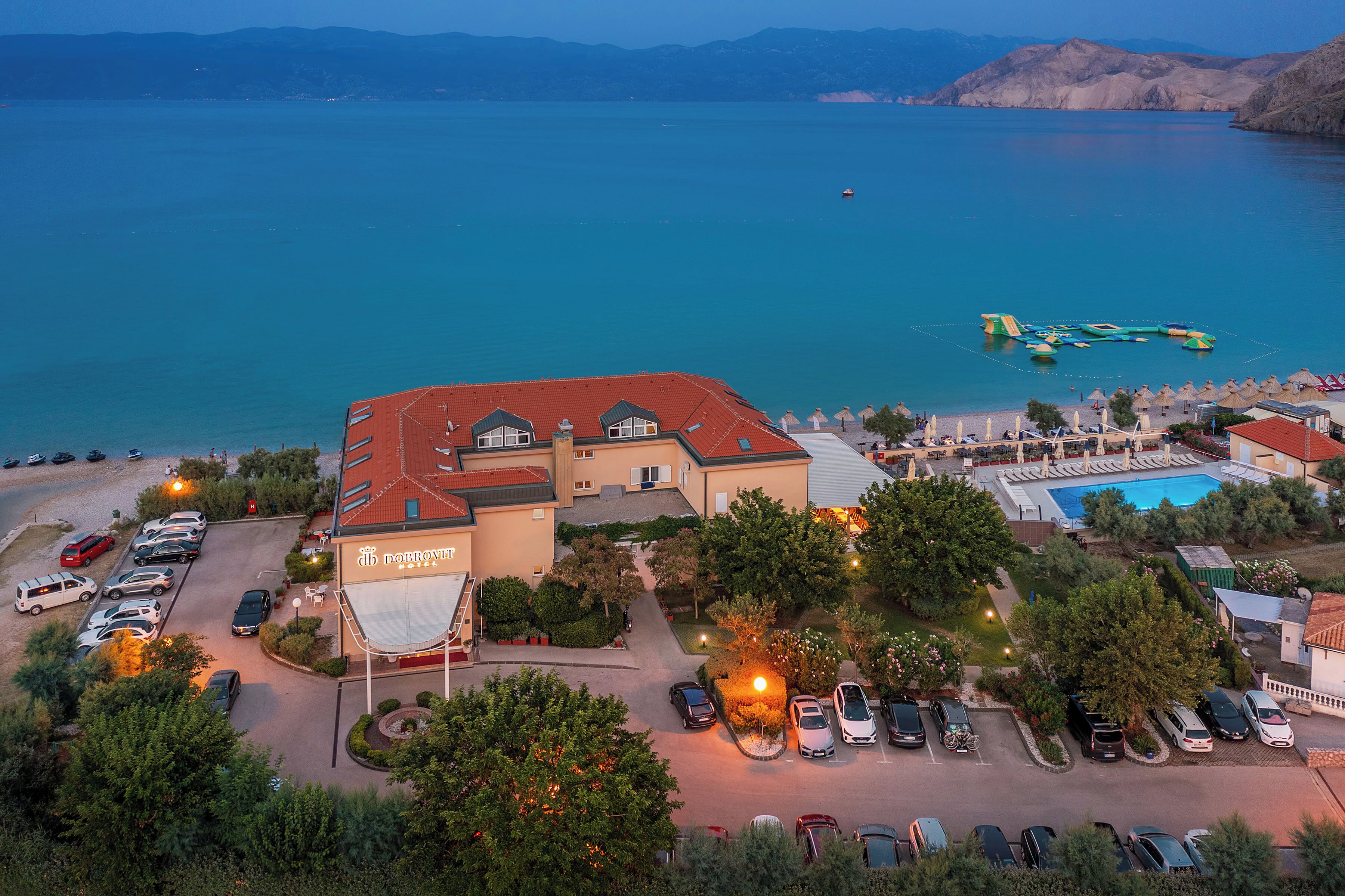 Un hotel con techo rojo, piscina y zona de playa con sombrillas.