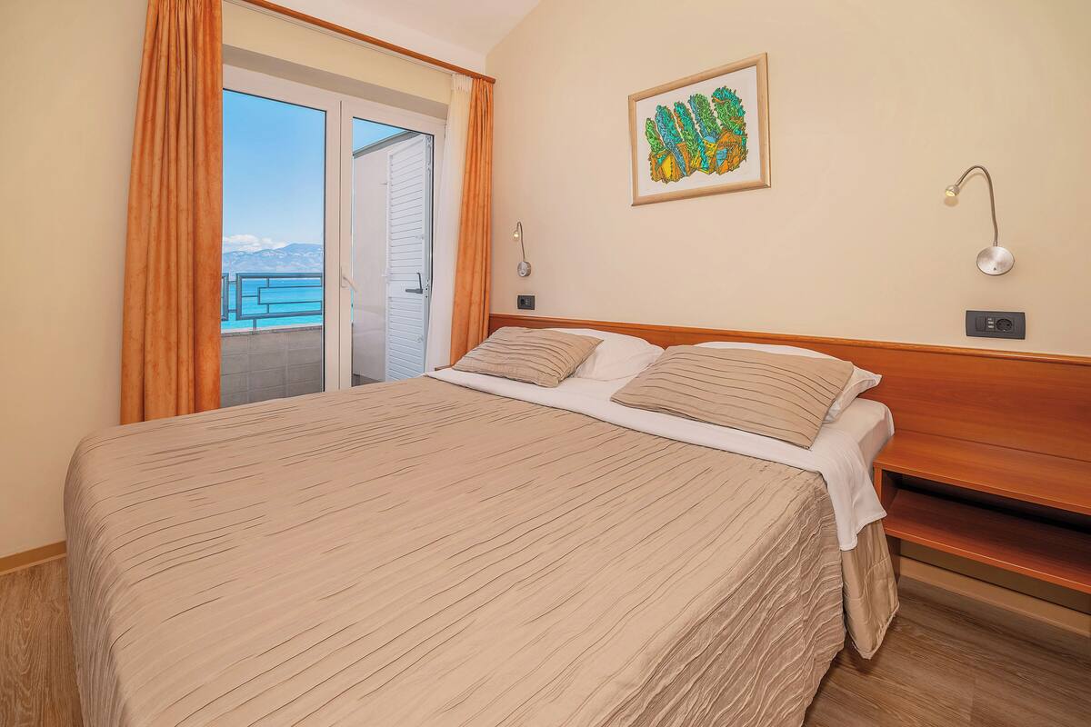A hotel room with a large bed, a sliding glass door to a balcony, a wall-mounted lamp, and a framed artwork.