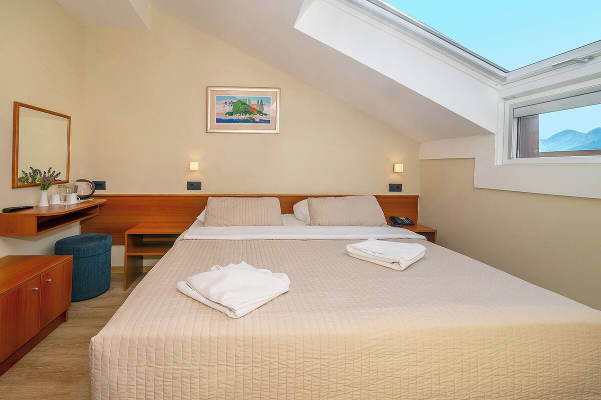 A neatly arranged hotel room with a large bed, a desk, a chair, a small ottoman, a mirror, and a painting on the wall.