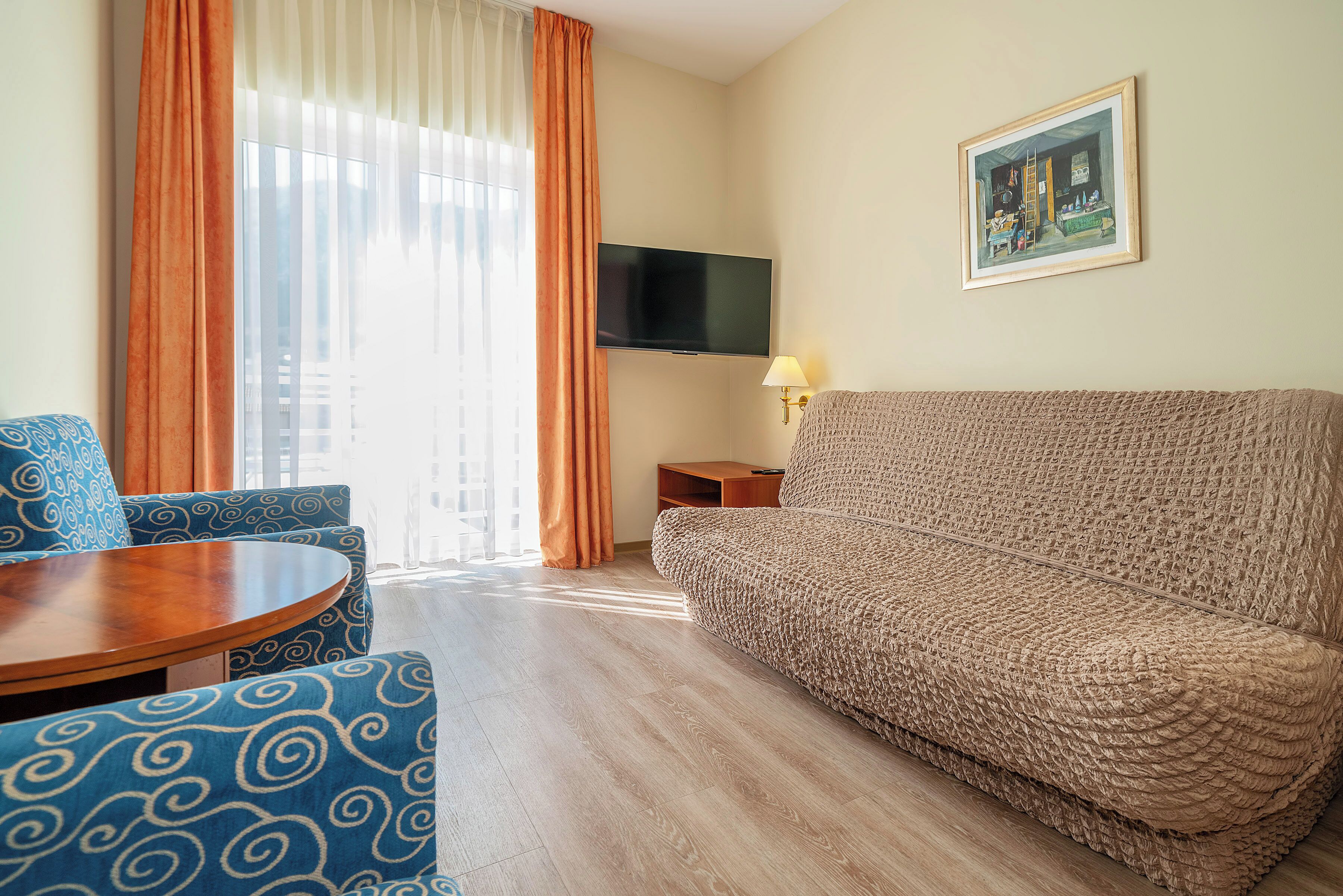 A hotel room with a sofa, armchair, TV, and a painting on the wall.