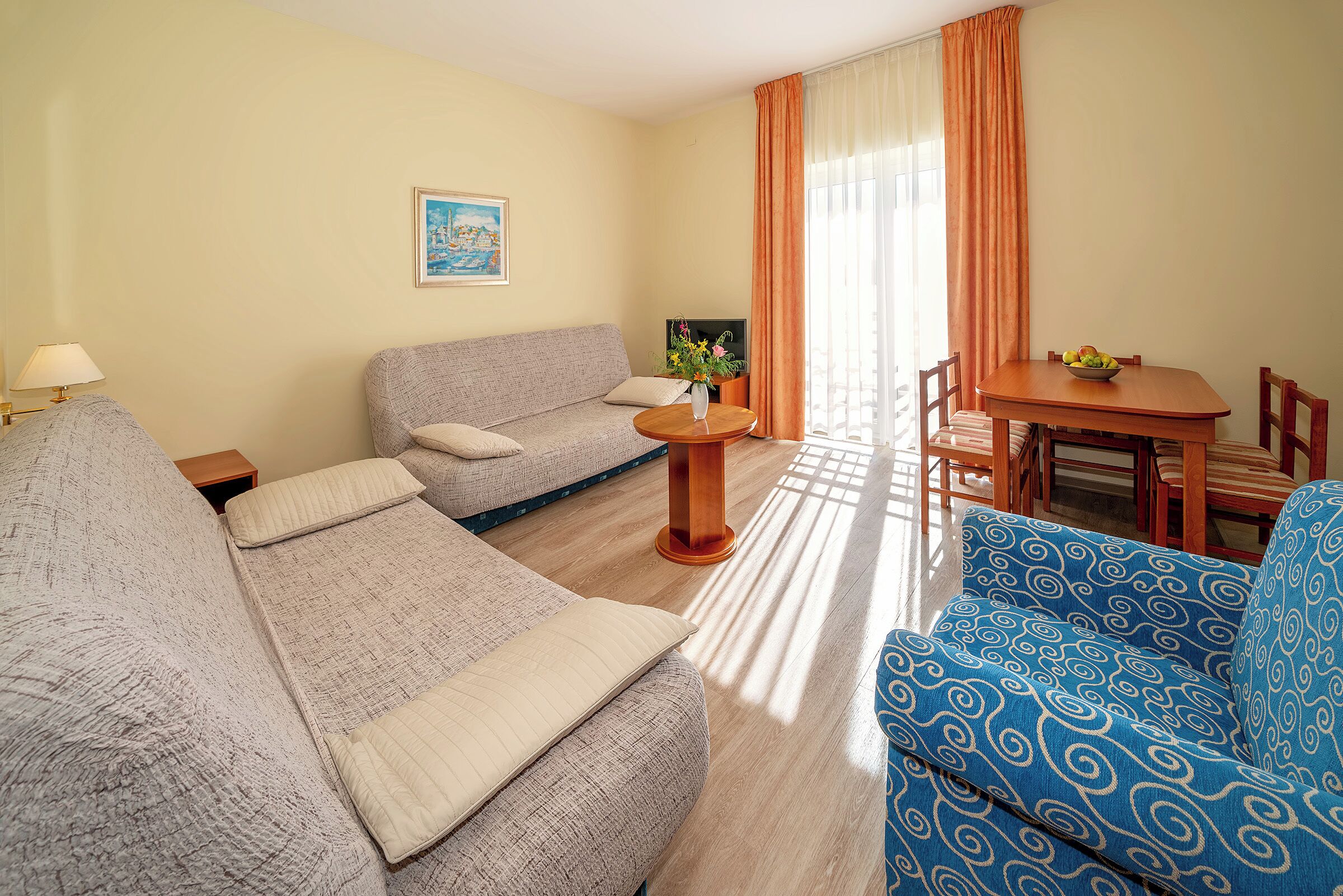 Una sala de estar con un sofá, un sillón azul estampado, una mesita con un frutero y una mesa de comedor con sillas.