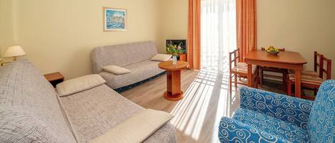 A living room with a sofa, a blue patterned chair, a small table with a bowl of fruit, and a dining table with chairs.