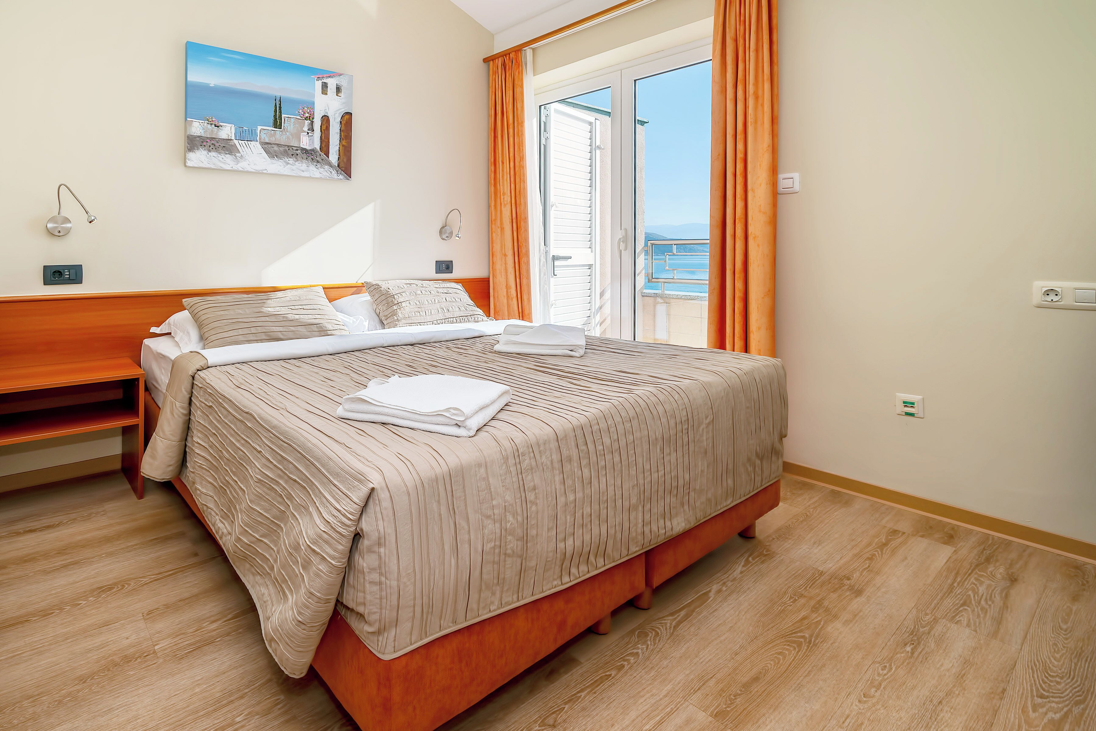 A neatly made bed with a striped bedspread and pillows, a wooden nightstand, a wall-mounted lamp, and a painting of a coastal scene.