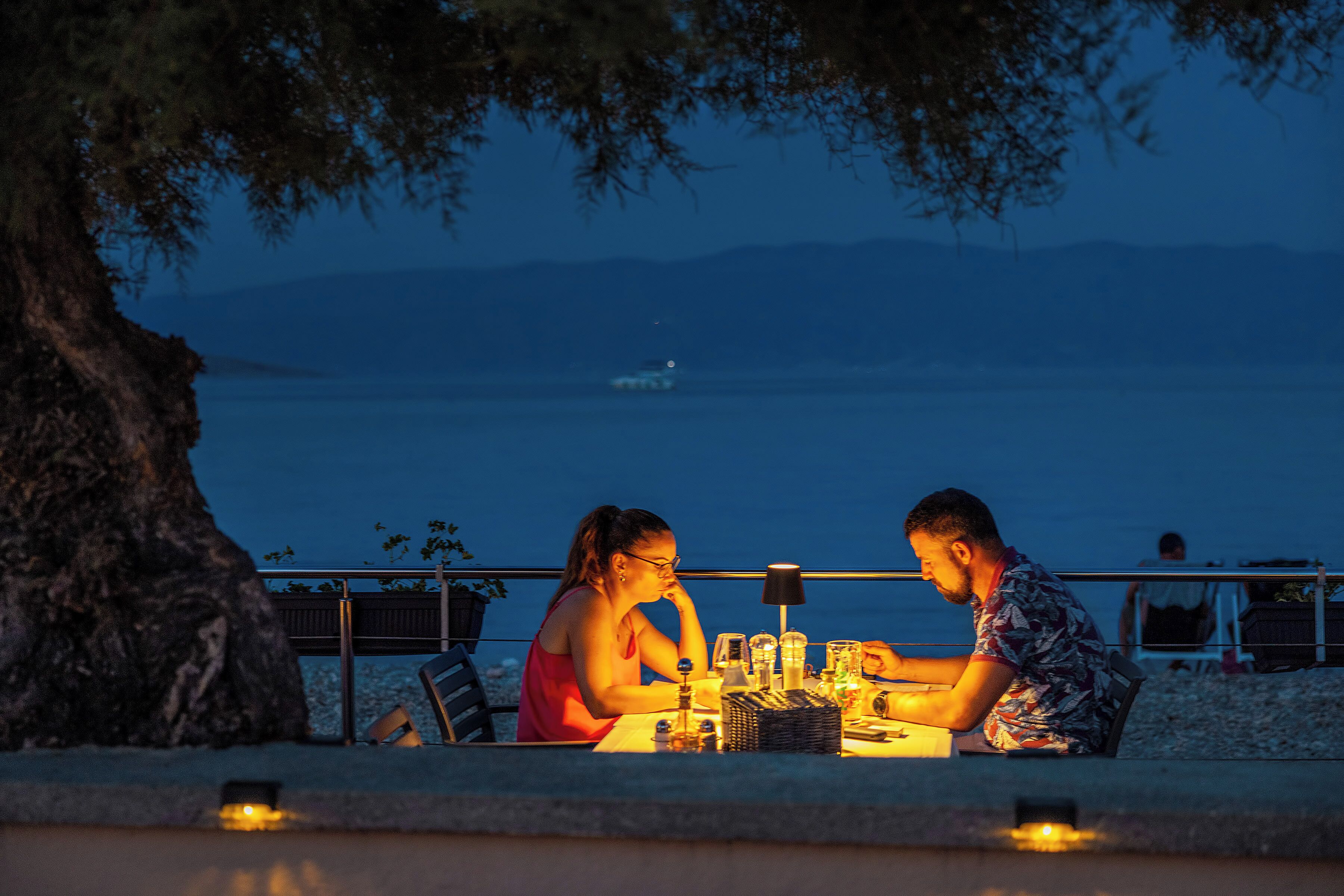 Un couple est assis à une table en plein air, profitant d’une vue sur la mer et les montagnes en arrière-plan.