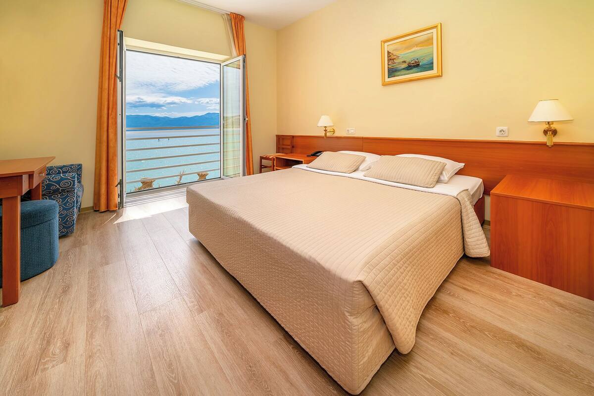 A hotel room with a large bed, a desk, a chair, and a view of the sea through a sliding door.