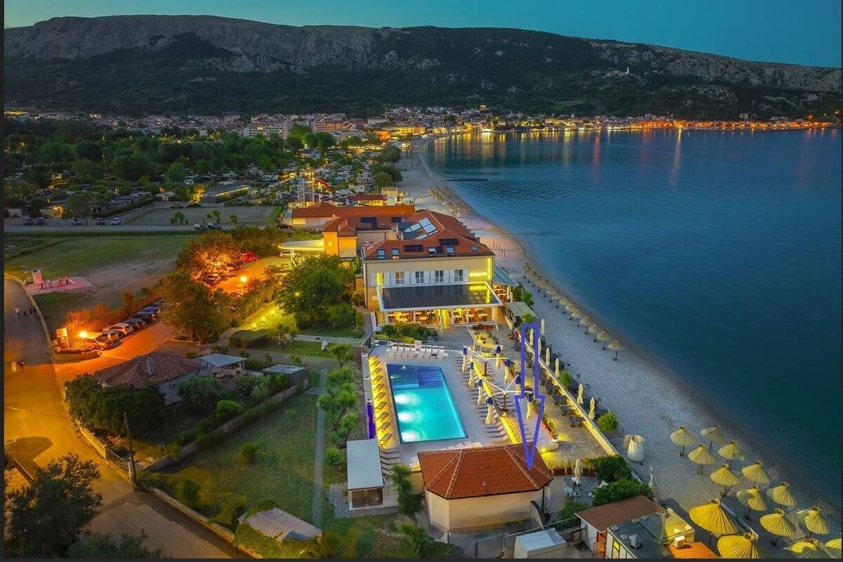 A coastal hotel with a swimming pool, beach umbrellas, and a view of the sea and mountains.