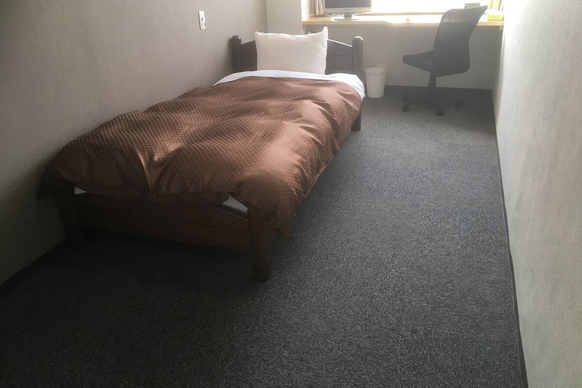 Een kleine eenpersoonskamer met een bureau en stoel, vloerbedekking en een raam voor natuurlijk licht.