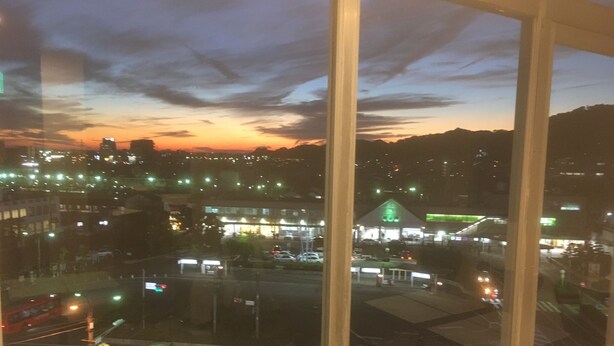 Une vue depuis une fenêtre donnant sur un paysage urbain au crépuscule, avec des bâtiments illuminés et un ciel dégagé.