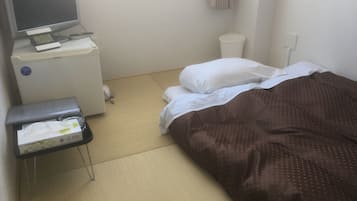 Habitación de hotel con una cama, una mesita de noche con televisor y despertador, un escritorio pequeño con computadora y un cesto de basura.