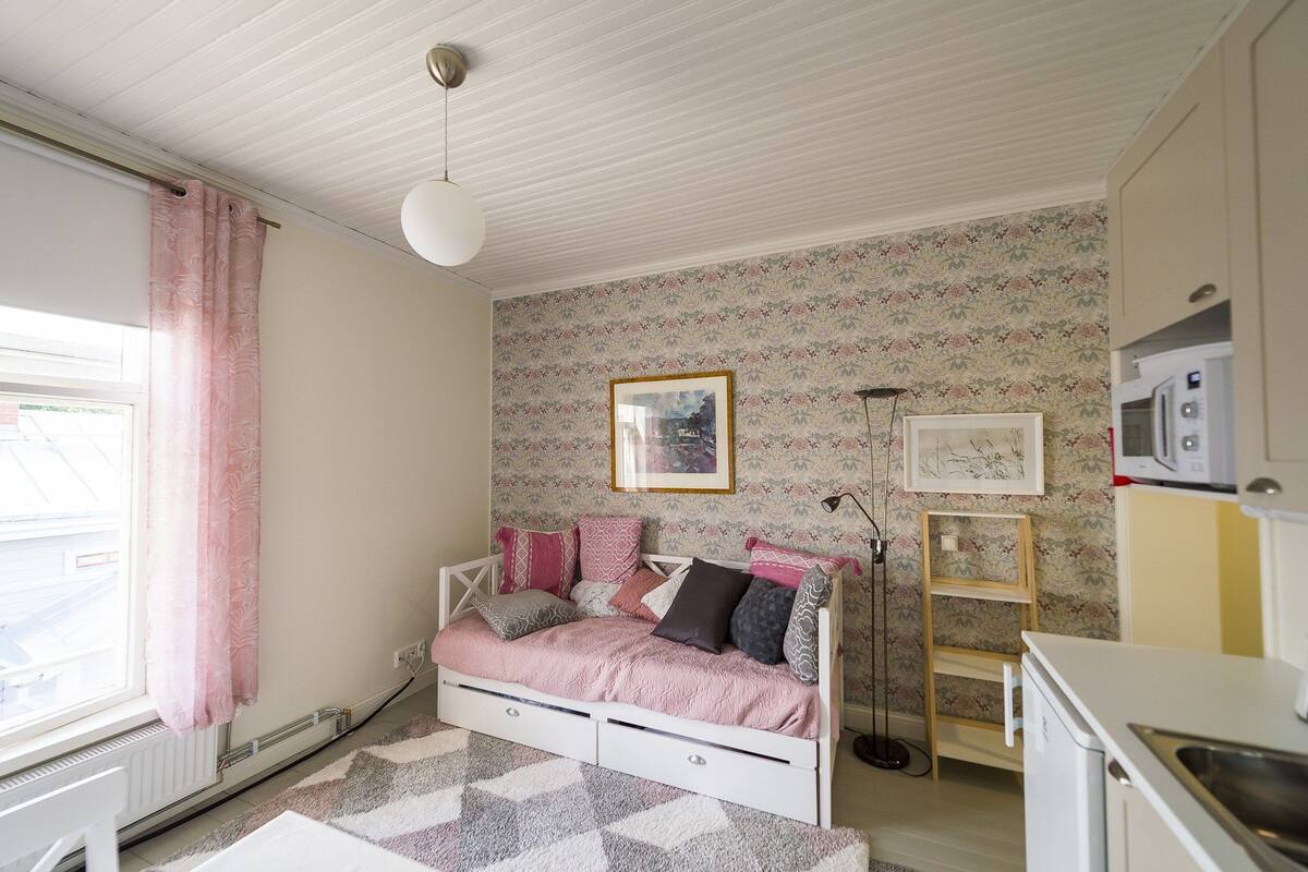 A bedroom with a white bed, pink bedding, a small desk, and a kitchenette.