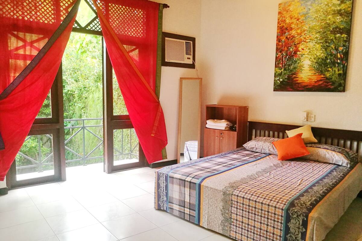 A bedroom with a bed, a wardrobe, a window with red curtains, and a painting on the wall.