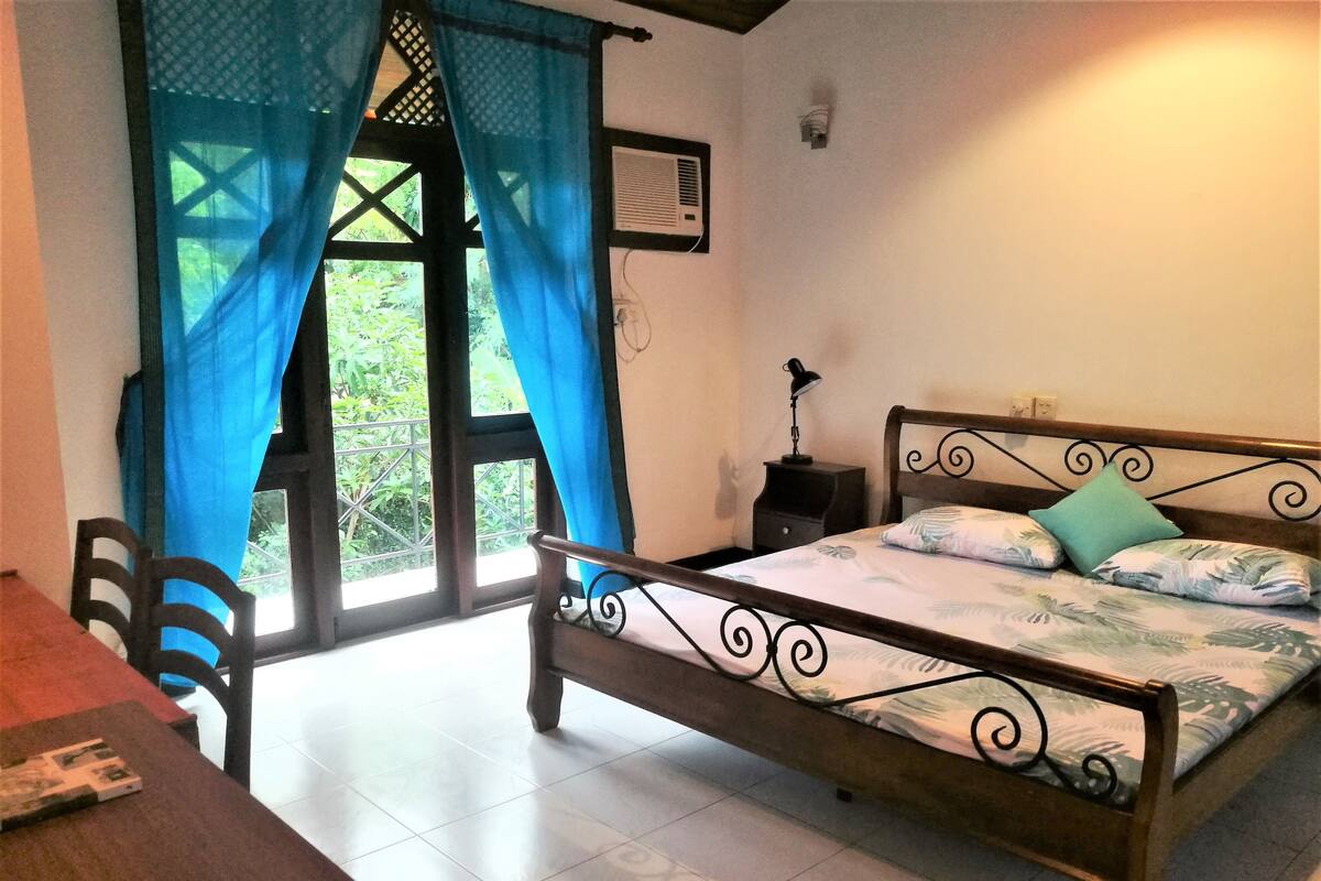 A bedroom with a wooden bed, a desk, and a chair. There is a window with blue curtains and a view of greenery outside.