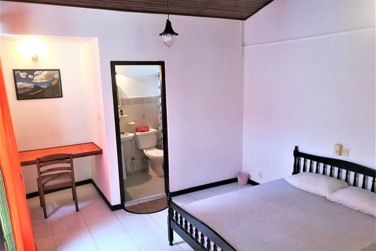 A bedroom with a bed, a desk, a chair, a bathroom visible through a door, and a hanging light fixture.