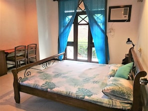A single bed with a floral bedspread, a nightstand with a lamp, a chair, and a window with blue curtains. - Bhakthi Yoga Sri Lanka (Colombo)