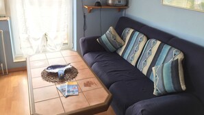 Un salon avec un canapé bleu foncé, des coussins à motifs, une table basse ornée d’un objet décoratif et d’un livre, ainsi que des étagères en bois où sont rangés des livres et une radio.