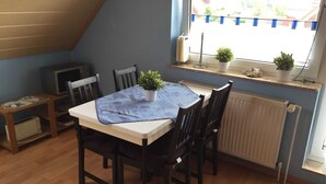 Un coin repas comprenant une table, des chaises, une plante en pot, un radiateur et une fenêtre donnant sur des maisons.