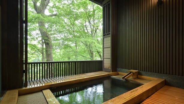 Un onsen japonés tradicional con una terraza de madera, un pequeño baño termal y vistas a árboles verdes exuberantes a través de un gran ventanal.