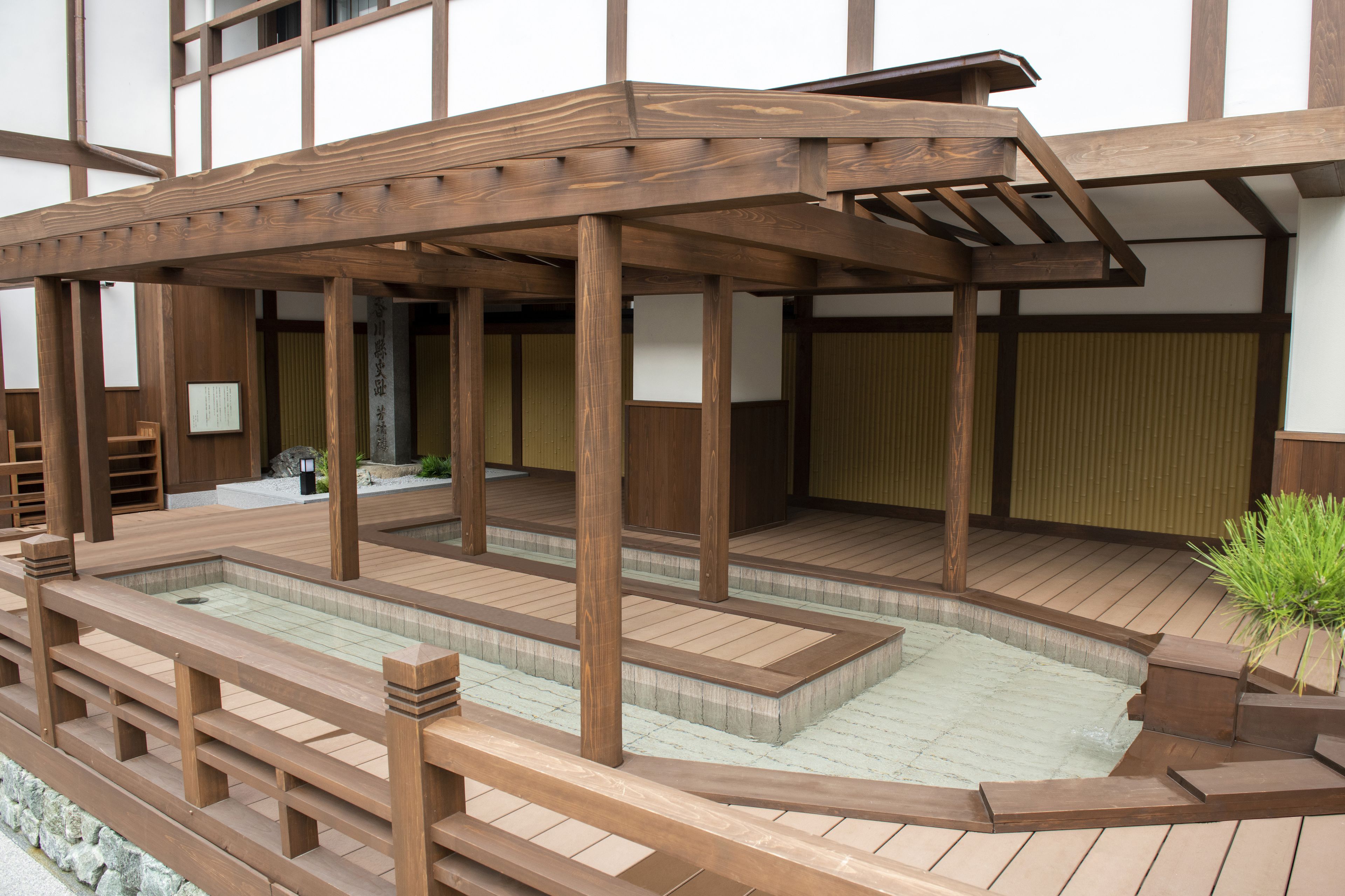 Una terraza tradicional japonesa de madera con un pequeño área de aguas termales, barandillas de madera y un sendero de piedra.