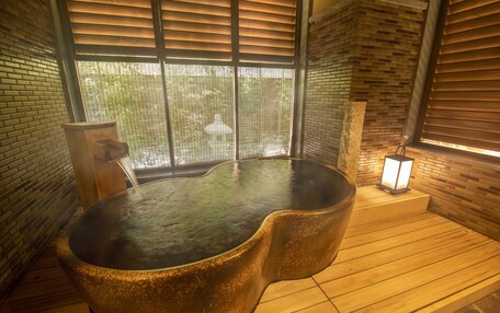 A hot tub in a room with wooden flooring and brick walls, a window with blinds, and a small table with a lamp.