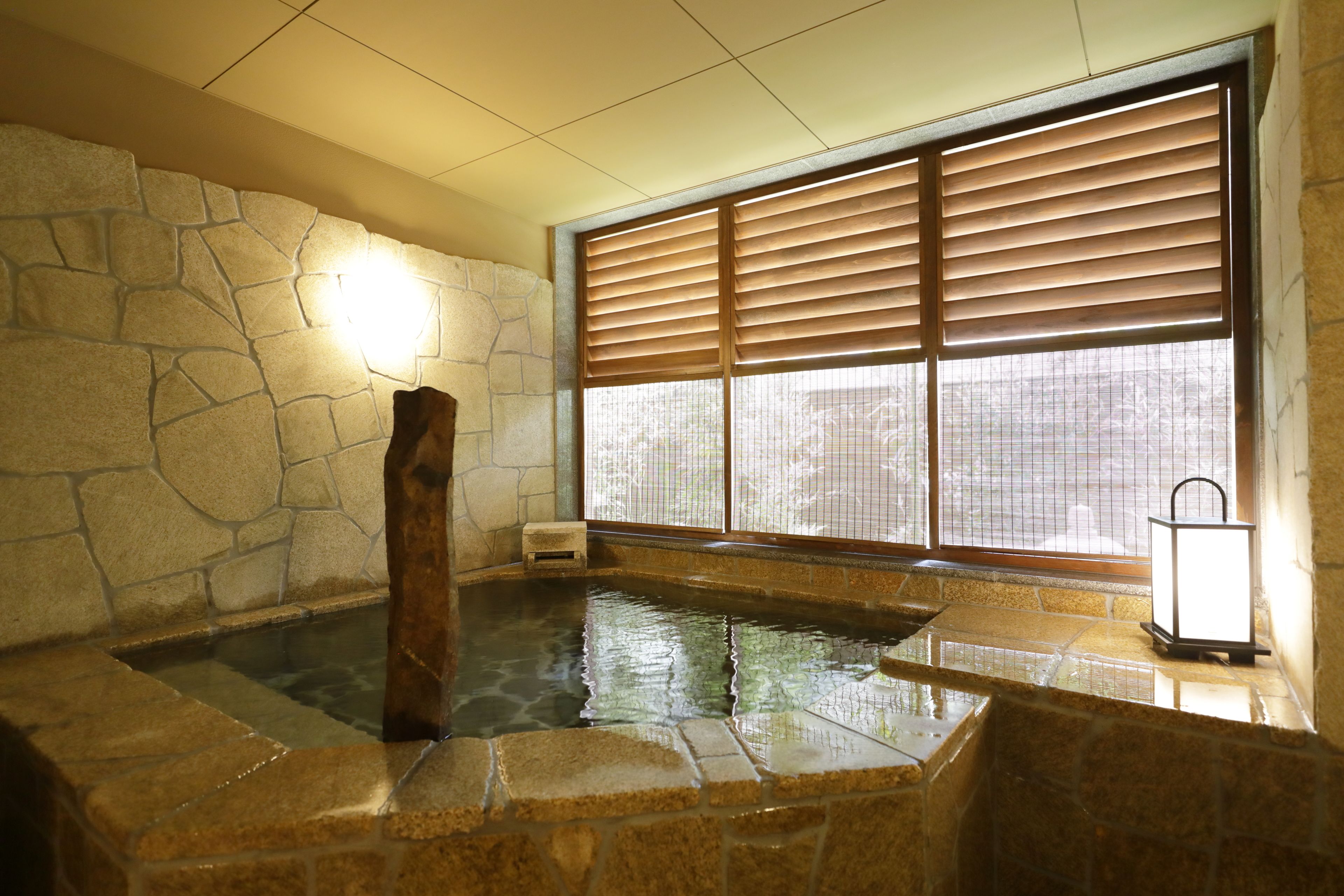 A stone-walled hot tub area with a wooden bench, a lantern, and a window with blinds.