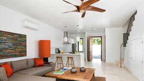 A modern living room with a sofa, coffee table, and a ceiling fan. There is a kitchen area in the background with a bar and stools. - Sugar Kube Villa Turks & Caicos – Romantic 1BR Villa with Private Pool & Rooftop Deck (Grace Bay)