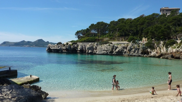 A beach with clear turquoise water, rocky cliffs, and a building on the hillside. - Apartment Can Serol 2 (Capdepera)