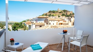 A balcony with white furniture, a small table with a bowl of fruit, and a view of a hilltop town with historic buildings. - Apartment Can Serol 2 (Capdepera)