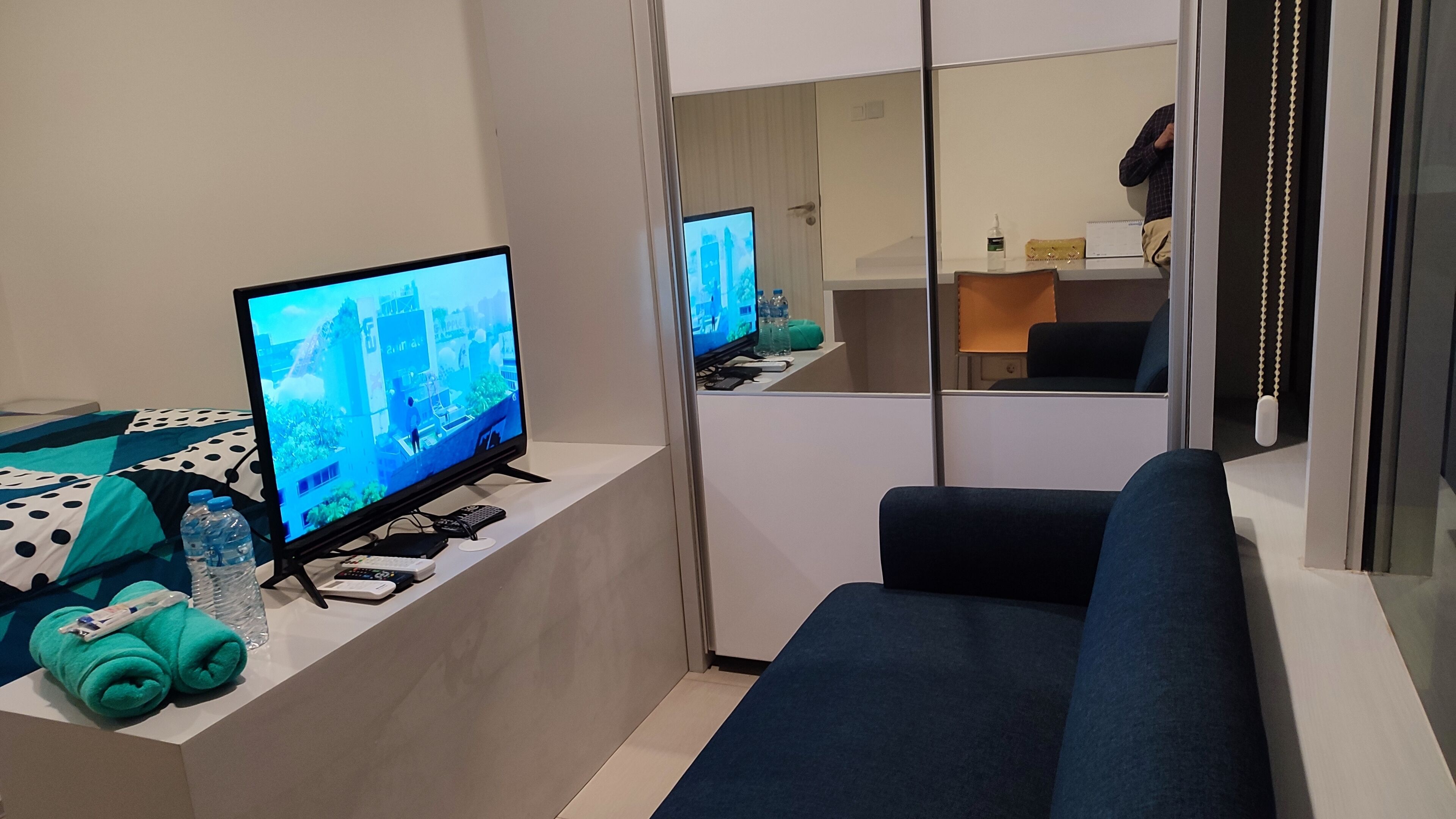Ein modernes Hotelzimmer mit großem Flachbildfernseher, einer Couch und einem Schreibtisch mit Computer und Fernbedienungen.