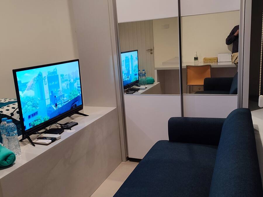 Ein modernes Hotelzimmer mit großem Flachbildfernseher, einer Couch und einem Schreibtisch mit Computer und Fernbedienungen.