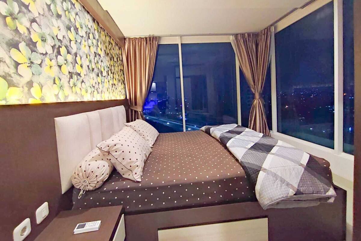 A bedroom with a large bed, a nightstand, and a view of the city at night.