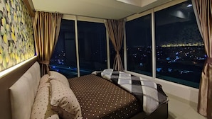 A bedroom with a large bed, multiple windows, and a city view at night. - Grand Kamala Lagoon By Mitra Propertindo (Bekasi)
