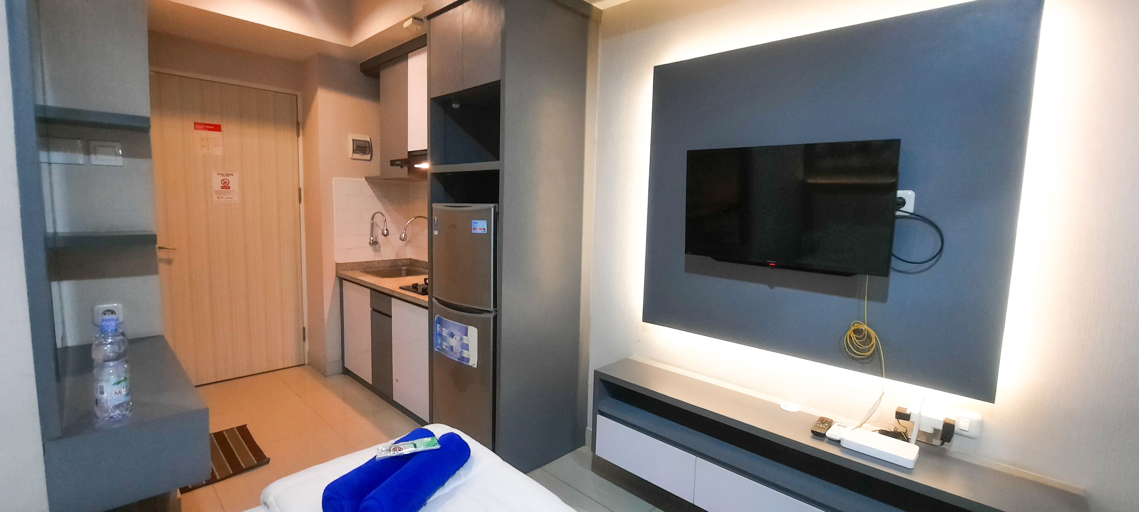 A modern hotel room with a kitchenette, a flat-screen TV mounted on the wall, and a bedside table with a blue towel and a bottle of water.