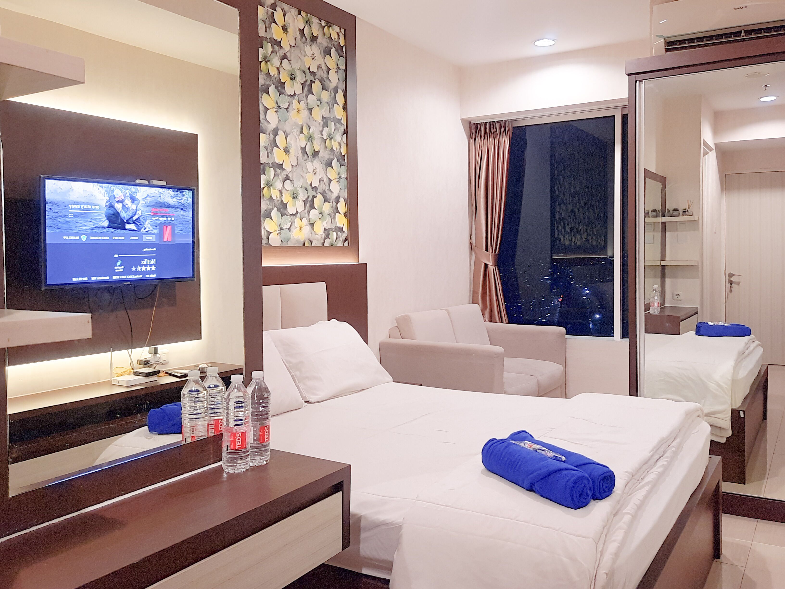 Ein Hotelzimmer mit einem Bett, einem Fernseher, einem Schreibtisch mit Wasserflaschen, einem Spiegel und einem Fenster mit Vorhängen.