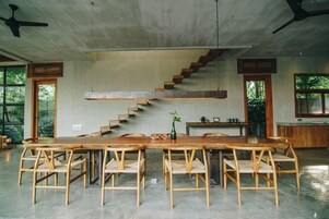 Una habitación amplia con una larga mesa de madera y sillas, una escalera y ventanas grandes.