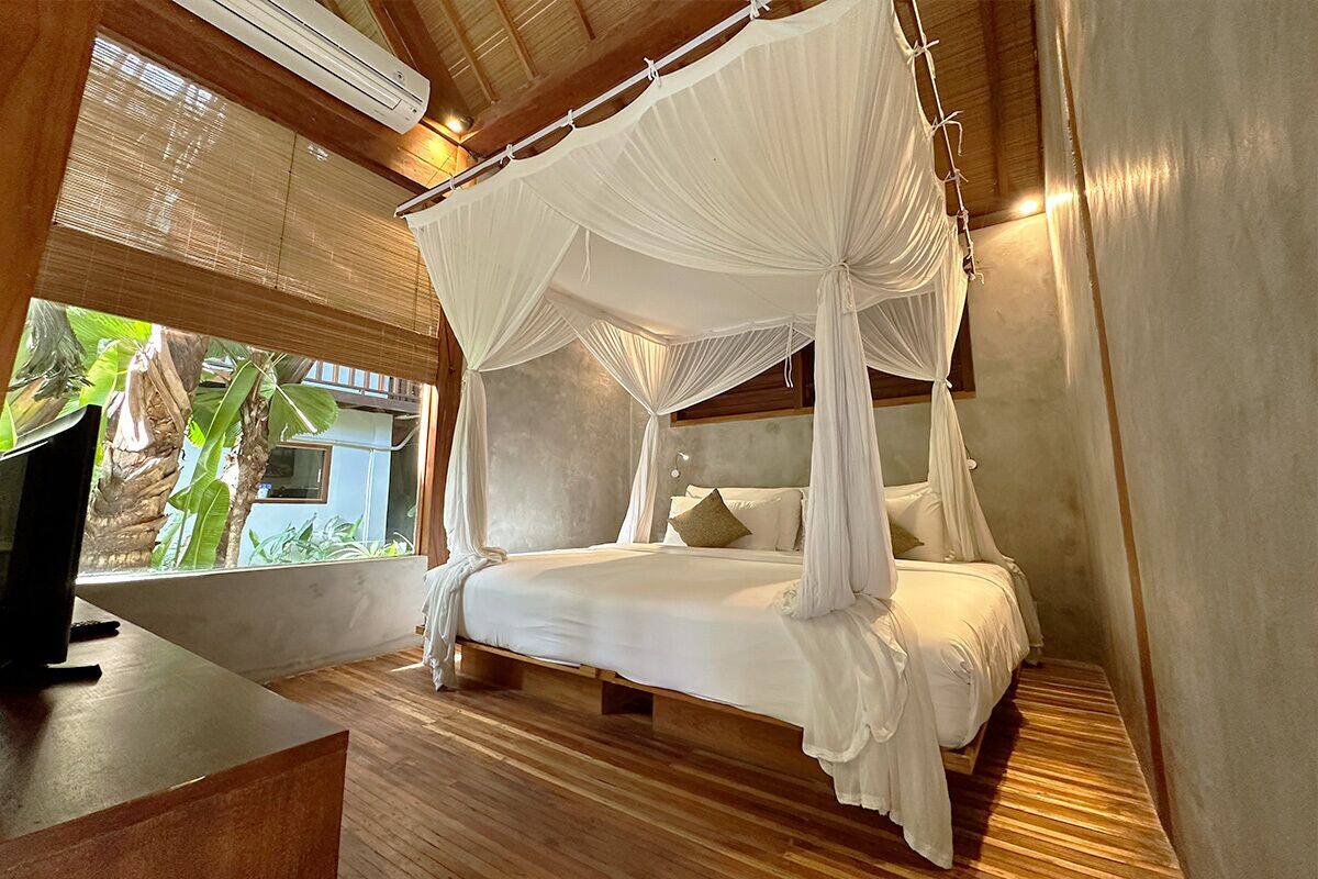 A four-poster canopy bed with white bedding, a wooden floor, and a view of tropical plants through a window.