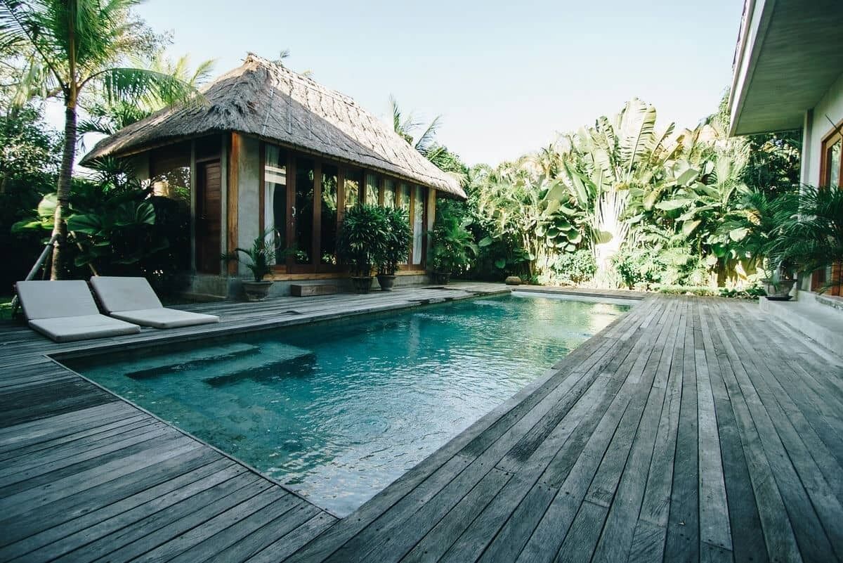 Una terraza de madera con piscina, sillas de descanso y una estructura con techo de paja, rodeada de vegetación tropical.