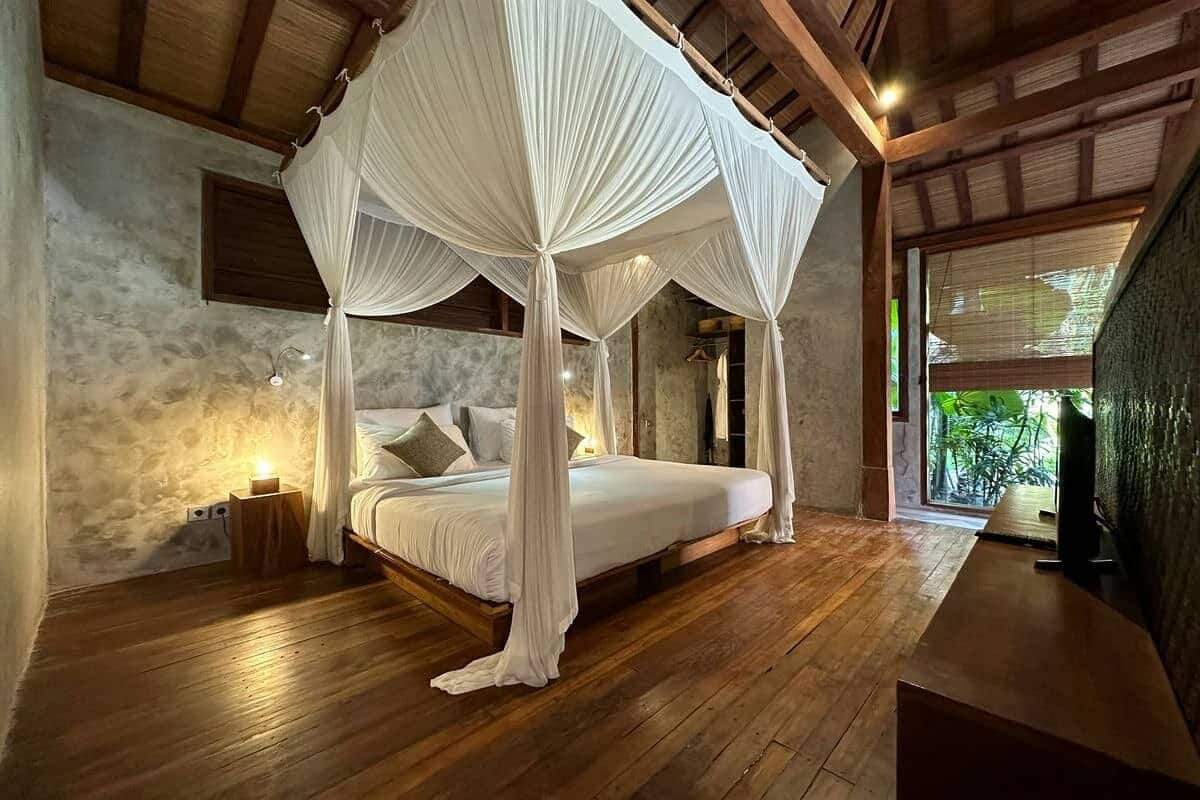 A four-poster canopy bed with a wooden floor, a nightstand with a lamp, and a view of greenery outside.