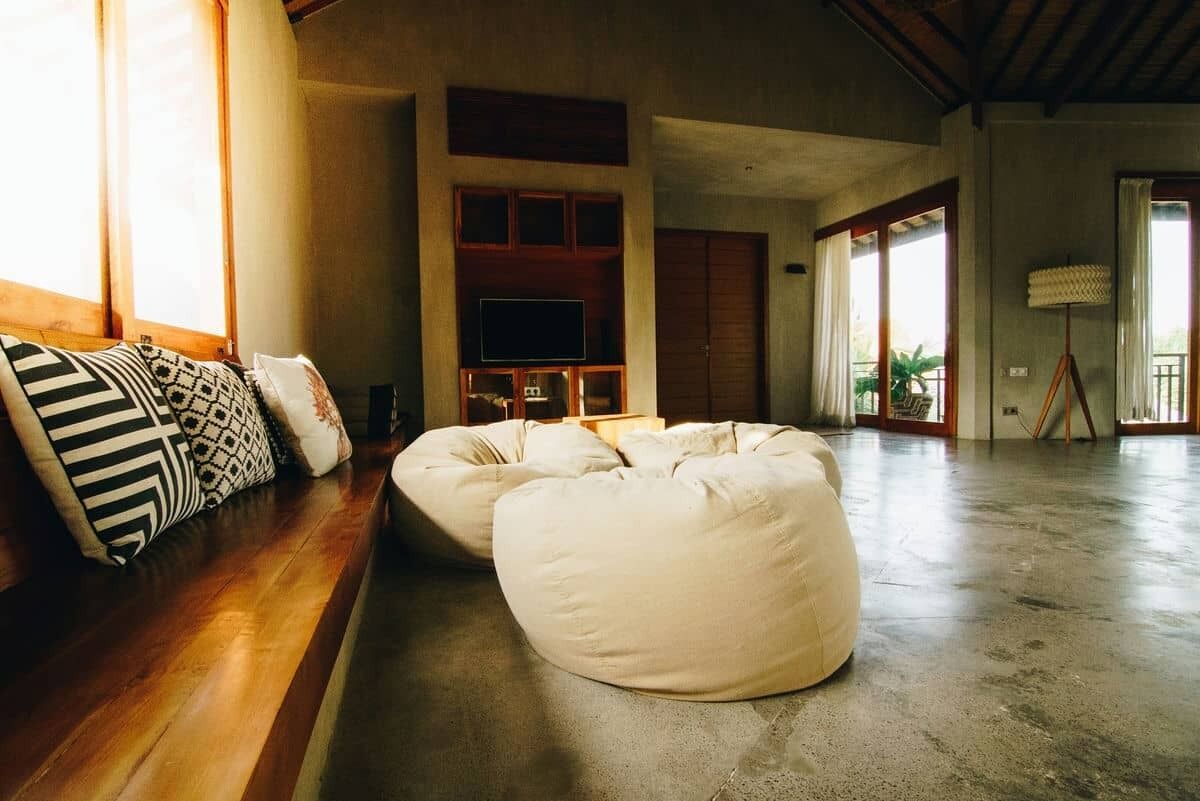 A spacious living room with a wooden bench, a beige bean bag, and a television on a wooden cabinet.