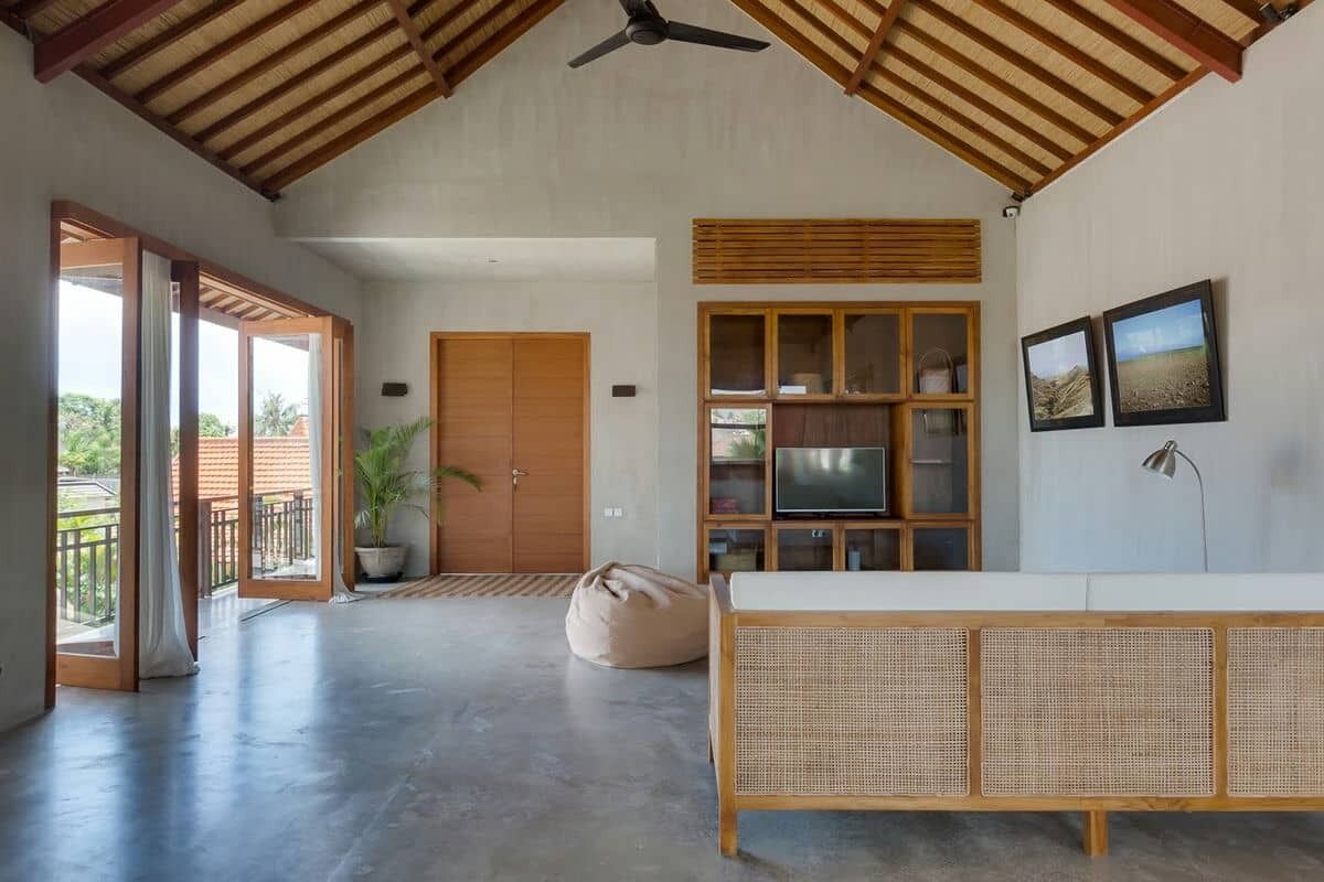 Una habitación amplia con techo alto, ventilador de techo, ventana grande, mueble de madera con televisor y un banco de ratán.
