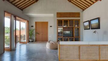 Una habitación amplia con techo alto, ventilador de techo, ventana grande, mueble de madera con televisor y un banco de ratán.