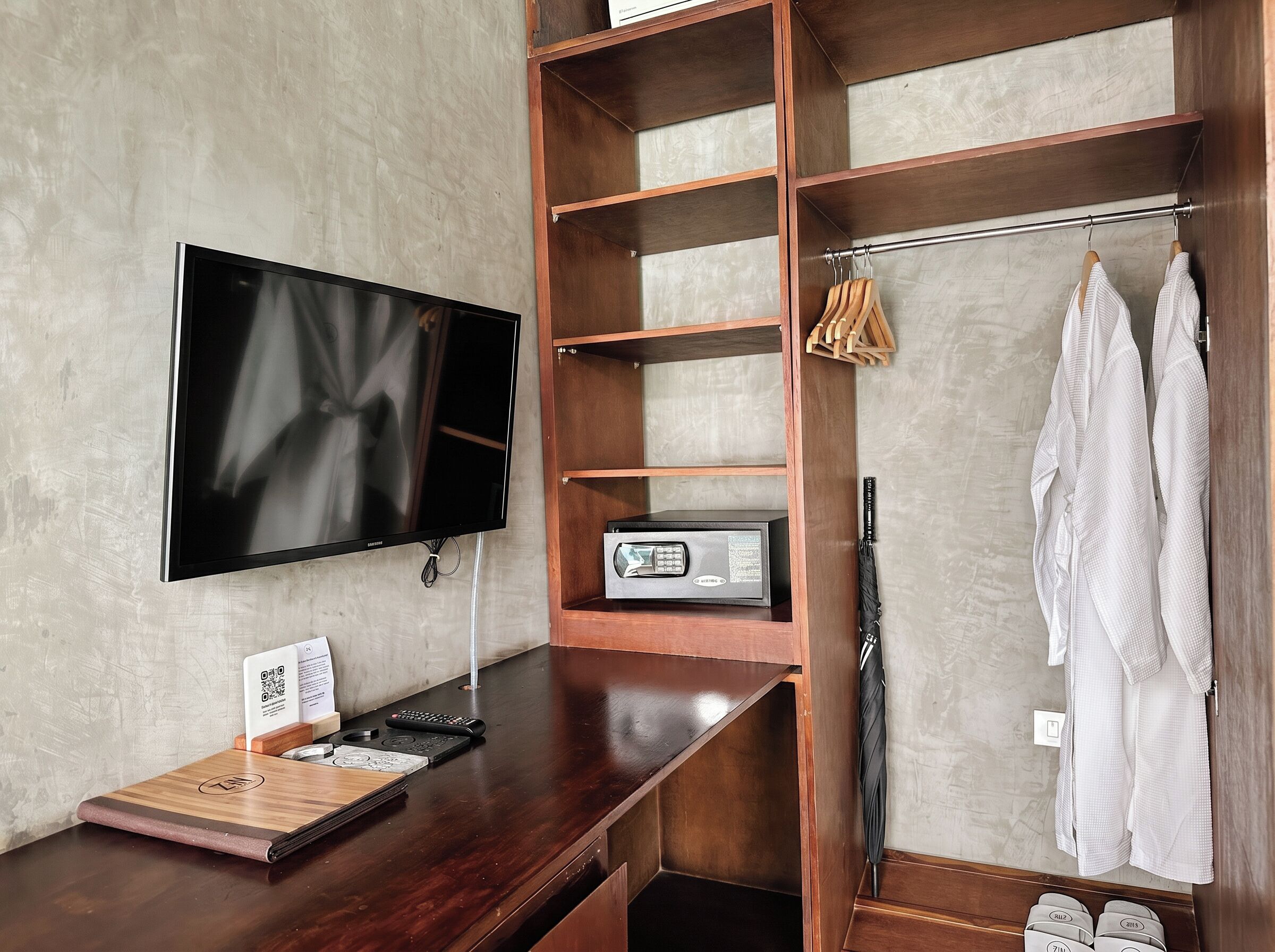 Habitación de hotel con televisión de pantalla plana, estanterías de madera, radio, perchero y dos albornoces blancos.